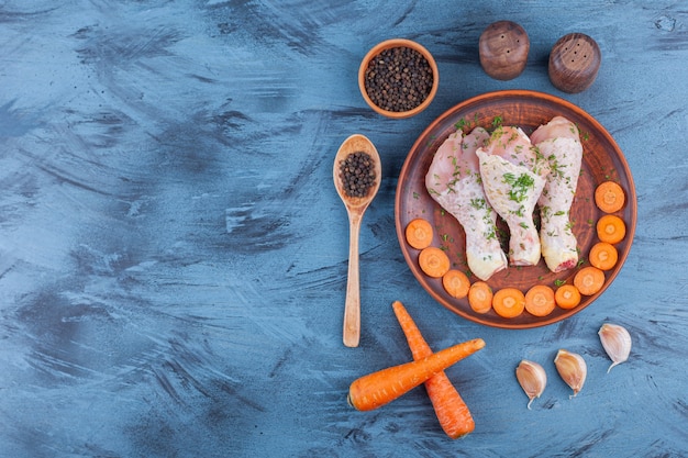 Pilons marinés et carottes tranchées sur une assiette en bois à côté d'épices, cuillère et ail, sur le fond bleu.