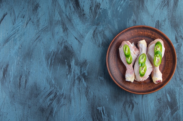 Pilons de poulet crus et poivrons tranchés sur une assiette, sur fond bleu.