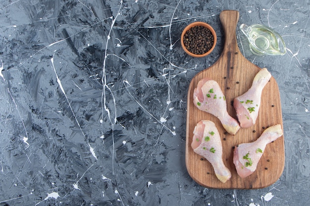 Pilons de poulet sur une planche à découper à côté de bols d'huile et d'épices , sur fond bleu.