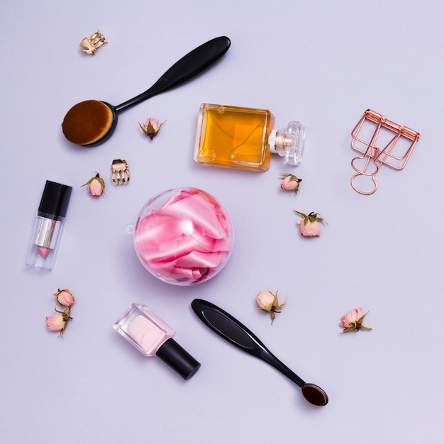 Photo gratuite pinceau de maquillage; rouge à lèvres; bouteille de parfum; vernis à ongles et embrayage avec des roses roses sur fond violet