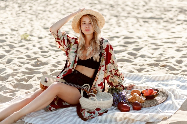 Pique-nique en campagne. Femme blonde romantique au chapeau de paille assis sur le couvercle sur la plage aux couleurs douces du coucher du soleil.
