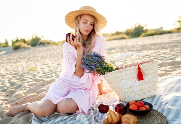 Pique-nique en campagne près de l'océan. Gracieuse jeune femme aux cheveux blonds ondulés dans une élégante robe rose, profitant des vacances et mangeant des fruits.