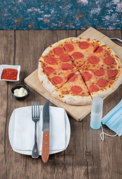 Pizza au pepperoni sur une planche en bois avec sauces, assiette, désinfectant pour les mains et masque autour