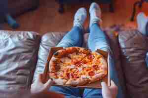 Photo gratuite une pizza dans une boîte sur les genoux d'un adolescent assis sur le canapé et regardant la télévision à plat, des aliments frais