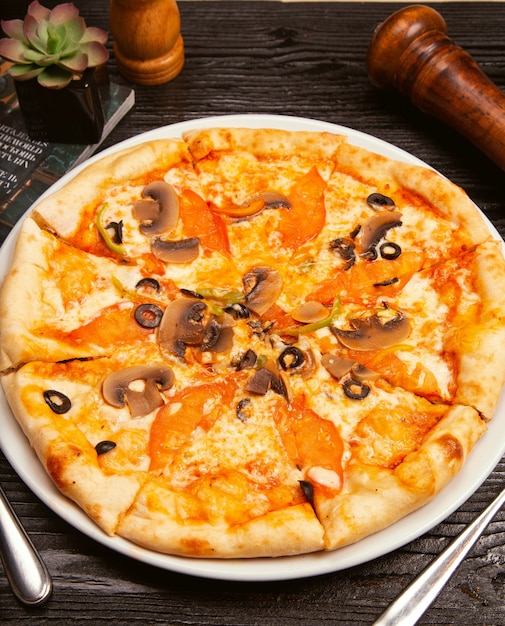 Pizza margarita aux olives noires, champignons, sauce tomate, tranches de tomate et fromage parmesan sur plaque blanche.