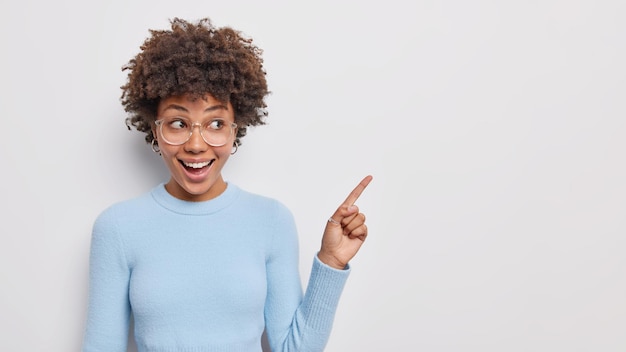 Place pour votre logo. Une belle jeune femme bouclée positive montre des points d'espace de copie dans le coin supérieur droit montre un bon produit porte un pull bleu isolé sur fond blanc.