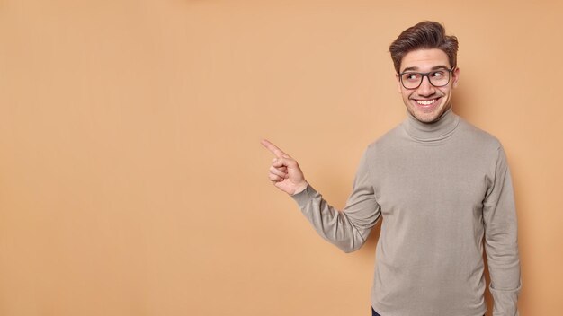 Placez votre annonce ici Beau jeune homme européen satisfait pointe sur un espace vide montre la place pour votre promo porte des lunettes transparentes et un col roulé isolé sur fond marron