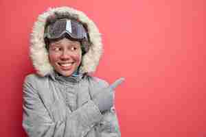 Photo gratuite plaisirs d'hiver et concept de ski. une femme afro-américaine à la peau sombre et heureuse en vêtements d'extérieur pointe sur un espace vide indiquant la direction vers le lieu de villégiature bénéficie de la saison de décembre.