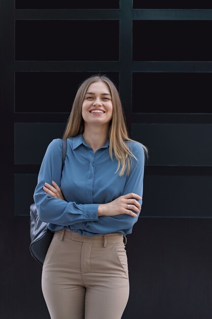 Plan de la belle jeune femme d'affaires portant une chemise en mousseline de soie bleue en position debout avec les bras croisés sur le mur noir