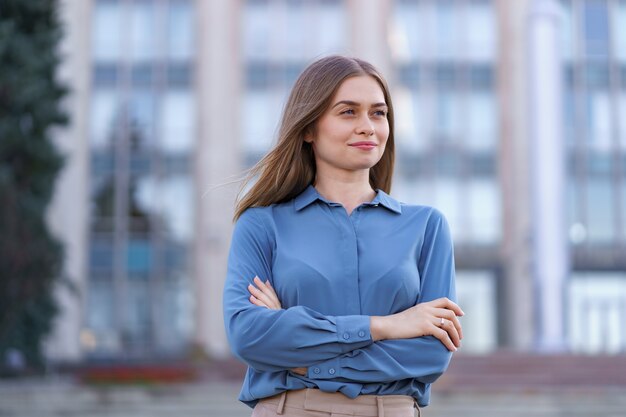 Plan de la belle jeune femme d'affaires portant une chemise en mousseline de soie bleue en se tenant debout sur le bâtiment dans la rue avec les bras croisés.