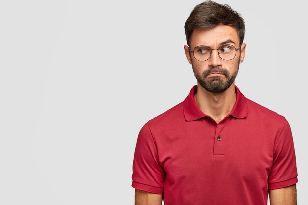 Plan candide d'un homme de race blanche perplexe avec un chaume sombre regarde de côté avec suspicion, lève les sourcils, porte un t-shirt rouge, remarque quelque chose sur l'espace vide. Concept de personnes et d'expressions faciales.