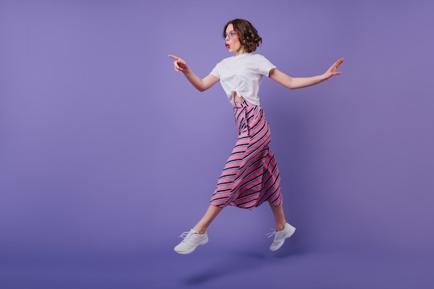 Plan complet d'une jolie fille excitée avec une coiffure ondulée sautant sur un mur violet. Élégante femme de bonne humeur en baskets s'amusant pendant la séance photo.