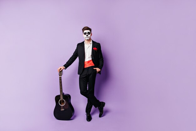 Plan complet d'un mec posant détendu avec une guitare. L'homme au visage peint en costume de style espagnol se penche sur la caméra.