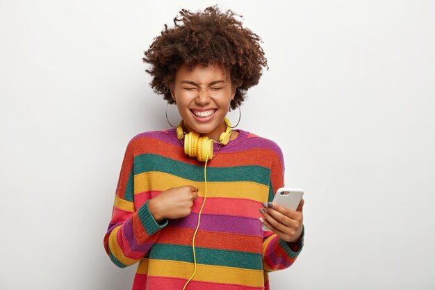 Plan demi-longueur d'une femme joyeuse ravie se pointe, tient un téléphone portable, exprime des émotions agréables, porte des boucles d'oreilles, un pull coloré, un casque autour du cou