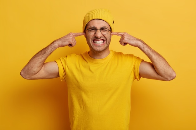 Photo gratuite plan demi-longueur d'un gars ennuyé serre les dents, bouche les oreilles avec l'index, ignore le son ennuyeux, ressent une douleur ou une douleur à l'oreille