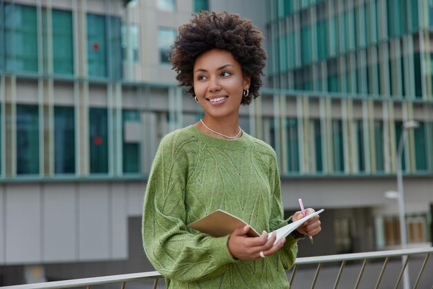 Plan extérieur d'une femme aux cheveux bouclés réfléchie se dresse contre un bâtiment urbain écrit des informations dans un cahier contient un stylo et des notes de bloc-notes texte crée un nouveau chapitre o livre vêtu d'un pull vert décontracté