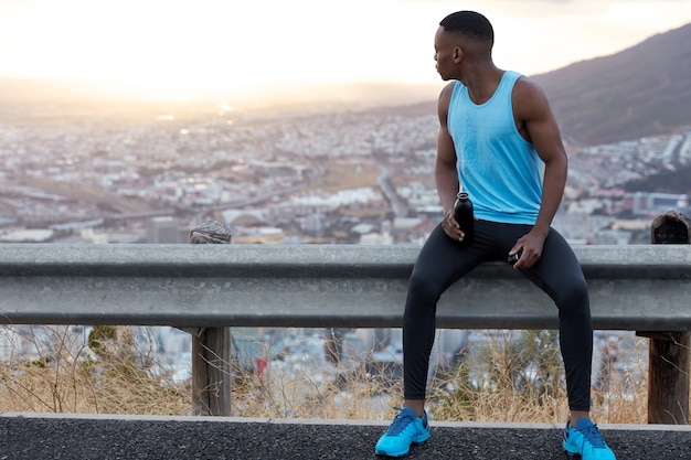 Plan extérieur d'un homme à la peau sombre concentré en arrière, porte des vêtements de sport décontractés, tient une bouteille d'eau froide, fait une pause au panneau de signalisation, des mannequins à la lumière du jour avec un espace libre pour votre promotion