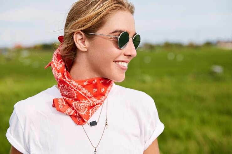 Photo gratuit plan extérieur d'une motocycliste joyeuse portant des lunettes de soleil élégantes et un bandana, apprécie la liberté en se promenant dans un champ vert calme, admire de magnifiques paysages naturels.