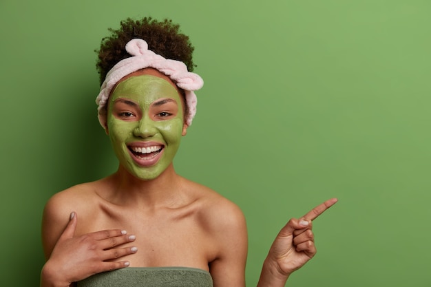 Plan d'une femme afro-américaine joyeuse à la recherche agréable applique un masque de beauté sur le visage, recommande un produit de cosmétologie, indique sur l'espace de copie, isolé sur un mur vert. Concept de soins de la peau