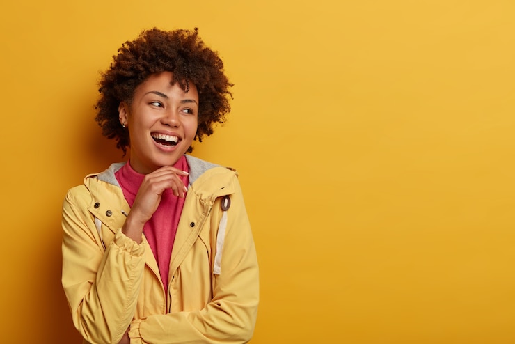 Photo gratuit plan d'une femme assez insouciante tient la main sous le menton, regarde de côté, porte une veste, pose contre un mur jaune, espace libre de côté pour votre contenu publicitaire. concept d'émotions et de sentiments