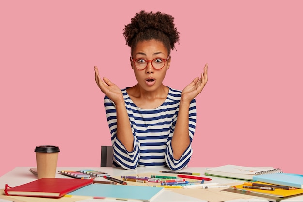 Photo gratuite plan d'une femme aux cheveux bouclés, terrifiée, impressionnée, garde les mains près du visage, regarde avec terreur, pose sur le lieu de travail, a une expression perplexe, travaille comme illustratrice, boit une boisson chaude, mannequins à l'intérieur