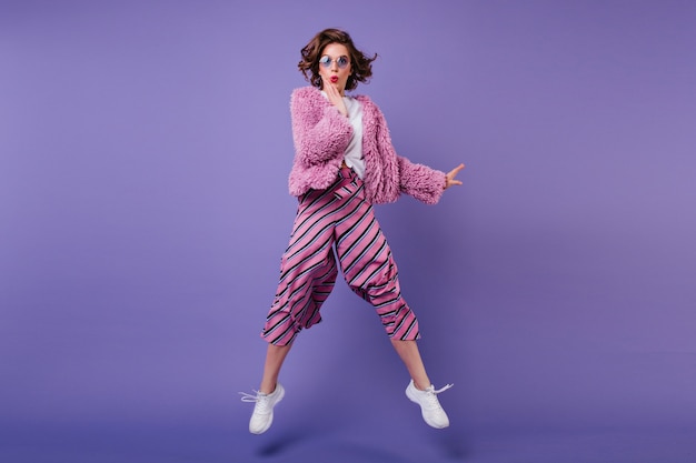 Plan d'une femme frisée heureuse en pantalon rayé sautant sur un mur violet. Portrait intérieur d'une fille merveilleuse à lunettes de soleil s'amuser.
