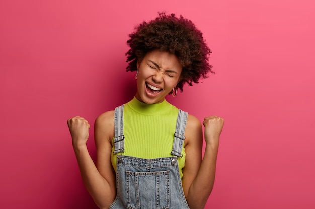 plan d'une femme frisée ravie se sent comme gagnante, serre les poings avec triomphe, porte des vêtements à la mode, s'exclame joyeusement, penche la tête, pose contre un mur rose, a des sentiments euphoriques