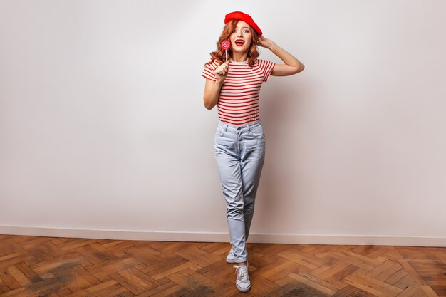 Plan d'une fille française en jeans à la mode mangeant une sucette. Photo intérieure d'une jolie femme au gingembre avec des bonbons.