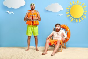 Photo gratuite plan d'un homme aux cheveux roux surpris avec de la crème solaire sur une peau brûlée par le soleil, se repose sur une chaise de plage, regarde avec choc la caméra, un ami se tient près de lui, porte un gilet de sauvetage et des lunettes de protection. deux compagnons sur la plage