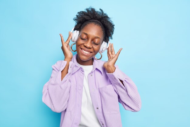 Plan horizontal d'une adolescente a la peau foncée, les cheveux bouclés naturels ferme les yeux avec plaisir incline la tête écoute de la musique avec des écouteurs