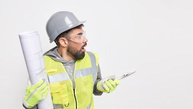 Plan horizontal d'un architecte masculin abasourdi détournant le regard avec une expression choquée tient un outil de construction et un plan vérifie le travail sur le chantier de construction pose contre la zone d'espace de copie de mur blanc