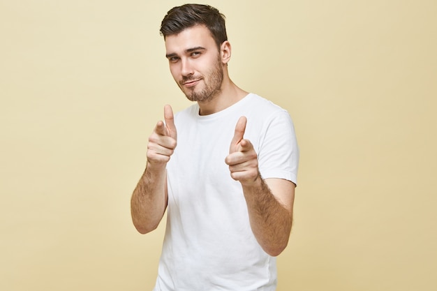 Plan horizontal d'un beau jeune homme charismatique aux cheveux noirs et chaume posant des doigts avant pointés isolés, ayant une expression faciale séduisante confiante, vous choisissant. Le langage du corps