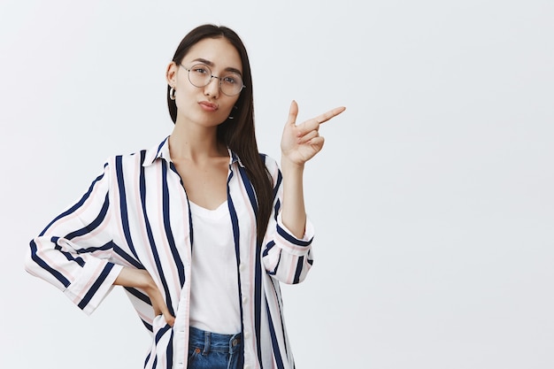 Plan horizontal de belle femme entrepreneur confiante et heureuse debout dans une pose féminine sûre d'elle-même sur un mur gris en tenue élégante, pointant vers la droite et pliant les lèvres mignonnes