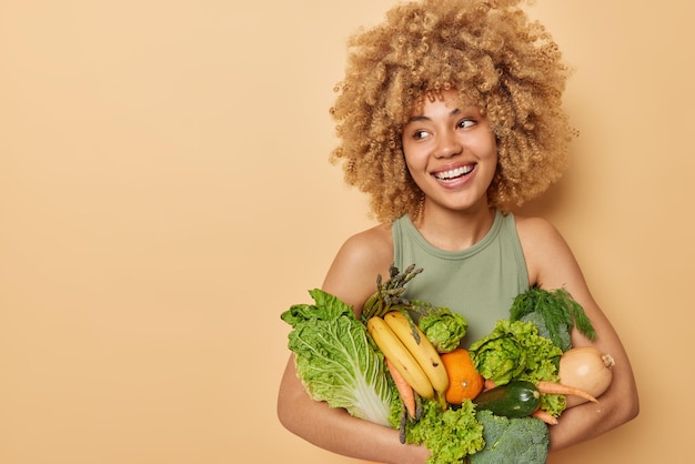 Plan horizontal d'une belle femme porte une variété de légumes et de fruits frais consomme une épicerie pleine de vitamines regarde au loin se dresse sur fond beige espace vide pour votre contenu promotionnel