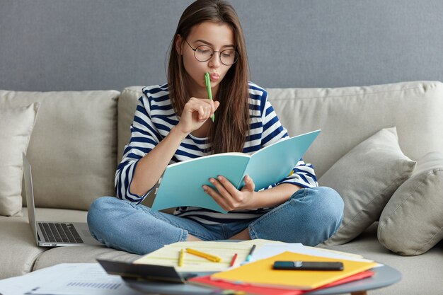 Plan horizontal d'une blogueuse occupée et réfléchie est assise sur un canapé avec un ordinateur portable, tient un stylo près de la bouche