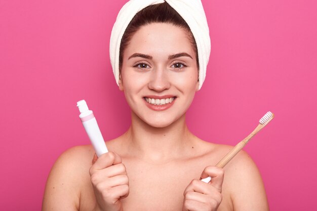 Plan horizontal d'une charmante femme souriante caucasienne isolée sur un mur de studio rose, prête à se brosser les dents, tenant du dentifrice et une brosse à dents dans les mains, a une peau douce et parfaite. Concept d'hygiène.