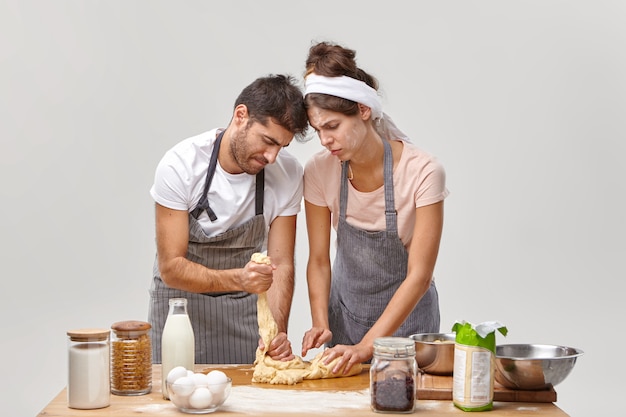 Photo gratuite plan horizontal d'un couple inexpérimenté prépare une pâte collante pour la première fois, étant de mauvais cuisiniers, regarder maladroitement, porter des tabliers, se tenir près de la table avec des produits. catastrophe de cuisine et échec de la cuisine