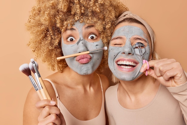 Plan horizontal de deux femmes subissent des procédures de beauté appliquer un masque d'argile utiliser des pinceaux cosmétiques et un recourbe-cils isolé sur fond marron préparer pour la fête Concept de cosmétologie des soins de la peau