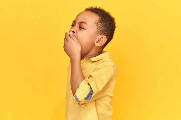 Plan horizontal d'un écolier africain endormi épuisé portant une chemise jaune couvrant la bouche avec la main bâillements d'être fatigué après une longue journée fatigante. Ennui, sommeil, heure du coucher et concept de literie