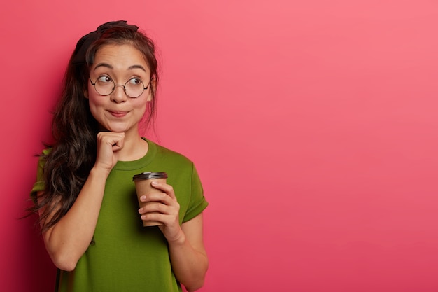 Plan horizontal d'une écolière asiatique songeuse rêve de vacances, tient le poing sous le menton, boit du café à emporter, apprécie la pause, se tient à l'intérieur contre un mur rose, porte des lunettes et un t-shirt.