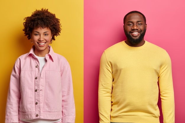 Photo gratuite plan horizontal d'une femme afro-américaine heureuse et d'un homme souriant satisfait, debout l'un à côté de l'autre contre un mur bicolore, heureux d'entendre d'excellentes nouvelles de l'interlocuteur, travailler en équipe.