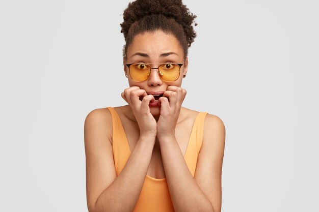 Plan horizontal d'une femme afro-américaine nerveuse en panique, mord les ongles, regarde anxieusement, porte des lunettes de soleil, pose contre le mur blanc. Concept de personnes, de sentiment et de réaction