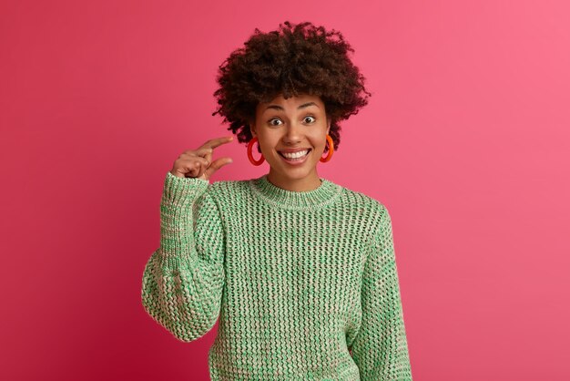 Plan horizontal d'une femme afro-américaine à la peau sombre montre un petit geste, mesure petit article, sourit agréablement, porte un pull tricoté, isolé sur un mur rose. Concept de langage corporel