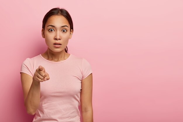 Plan horizontal d'une femme asiatique effrayée pointe l'index, se sent gênée de remarquer quelque chose d'étrange, a les cheveux noirs peignés en queue de cheval, porte un t-shirt rose, isolé sur un mur rose