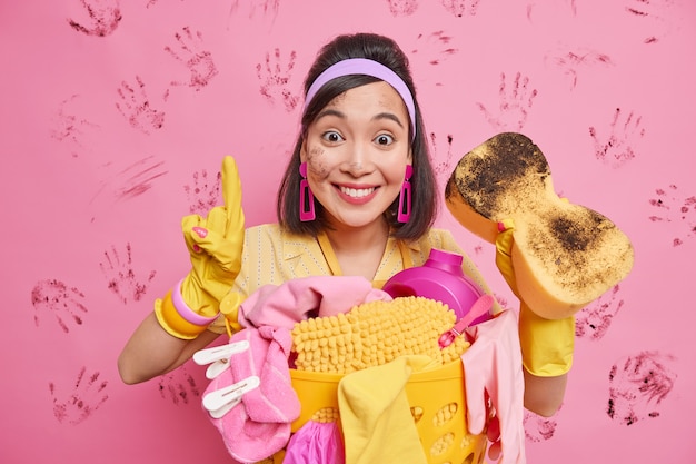 Plan horizontal d'une femme asiatique heureuse souriante garde agréablement le doigt levé montre les résultats de son travail tient une éponge ramasse le linge dans des poses de panier contre le mur rose