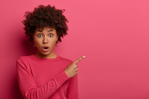 Photo gratuite plan horizontal d'une femme aux cheveux bouclés à la peau sombre et choquée garde la bouche ouverte, halète d'émerveillement, montre quelque chose à bas prix, porte un pull rose, fait des gestes à l'intérieur. concept de publicité