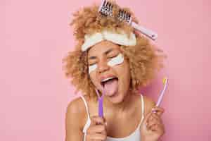 Photo gratuite plan horizontal d'une femme aux cheveux bouclés utilise des brosses à langue et une brosse à dents pour prendre soin de la cavité buccale applique des patchs de beauté porte un masque de sommeil et un t-shirt décontracté isolé sur fond rose