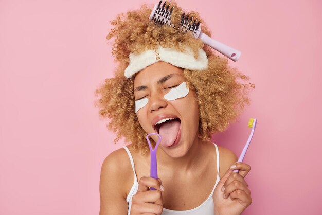 Plan horizontal d'une femme aux cheveux bouclés utilise des brosses à langue et une brosse à dents pour prendre soin de la cavité buccale applique des patchs de beauté porte un masque de sommeil et un t-shirt décontracté isolé sur fond rose