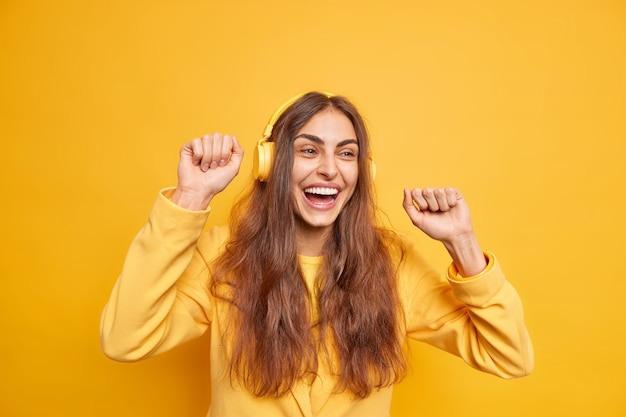 Plan horizontal d'une femme aux cheveux noirs optimiste utilise des écouteurs bouge le corps apprécie une nouvelle liste de lecture une expression joyeuse aime la mélodie porte une veste jaune sourit largement s'amuse