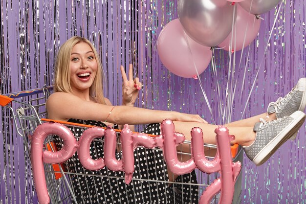 Plan horizontal d'une femme blonde heureuse dans un panier, fait un geste de paix, porte une robe et des chaussures de sport, s'amuse à la fête avec des ballons, isolé sur un mur violet. Concept de jour de fête